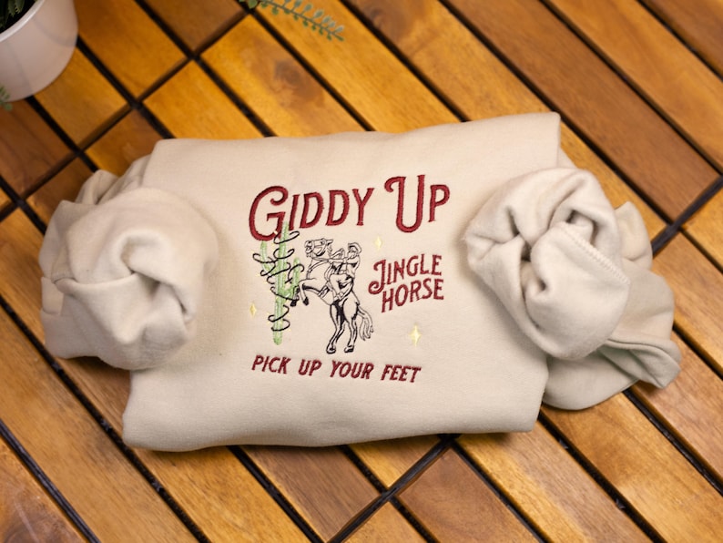 May include: A beige sweatshirt with the text "Giddy Up Jingle Horse Pick Up Your Feet" embroidered in red on the front.