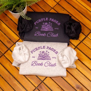 May include: A black sweatshirt and a light gray sweatshirt, each featuring a purple graphic of an open book with flowers and the text Purple Pages Book Club Est. 2024. Both garments are folded and include matching fabric scrunchies on the sleeves.