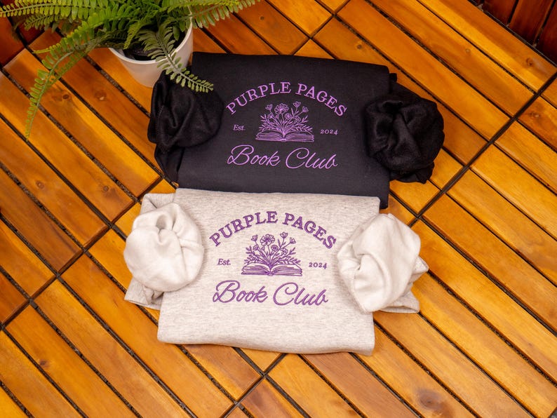 May include: A black sweatshirt and a light gray sweatshirt, each featuring a purple graphic of an open book with flowers and the text Purple Pages Book Club Est. 2024. Both garments are folded and include matching fabric scrunchies on the sleeves.