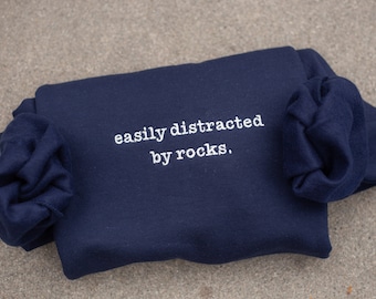 May include: A navy blue sweatshirt with the text "easily distracted by rocks." embroidered in white.