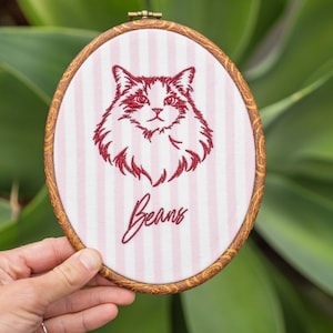 May include: An oval embroidery hoop with a wood-grain frame containing light pink and white vertical striped fabric. The fabric features a dark red embroidered portrait of a fluffy cat and the word Beans in a cursive script font below the cat.