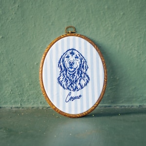 May include: An oval embroidery hoop frame with a wood-look finish containing white fabric with light blue vertical stripes. The fabric features a dark blue embroidered portrait of a dog and the name Cosmo written in cursive script below the dog.