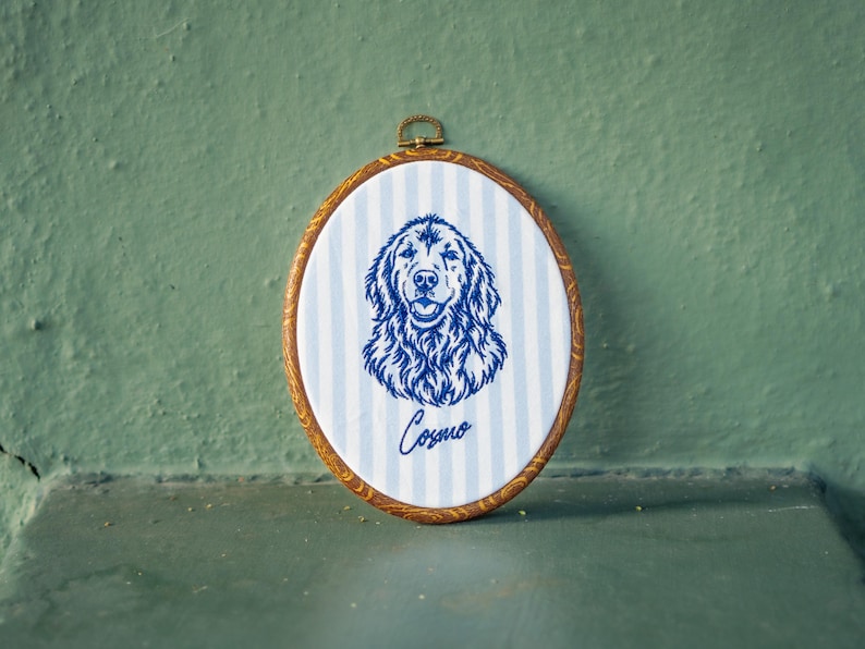 May include: An oval embroidery hoop frame with a wood-look finish containing white fabric with light blue vertical stripes. The fabric features a dark blue embroidered portrait of a dog and the name Cosmo written in cursive script below the dog.
