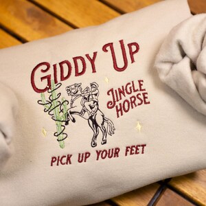 May include: A beige sweatshirt with the text "Giddy Up" in red embroidery. The design also includes a cartoon cowboy riding a horse with the text "Jingle Horse" and "Pick Up Your Feet" in red embroidery.