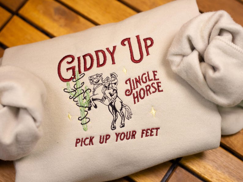 May include: A beige sweatshirt with the text "Giddy Up" in red embroidery. The design also includes a cartoon cowboy riding a horse with the text "Jingle Horse" and "Pick Up Your Feet" in red embroidery.