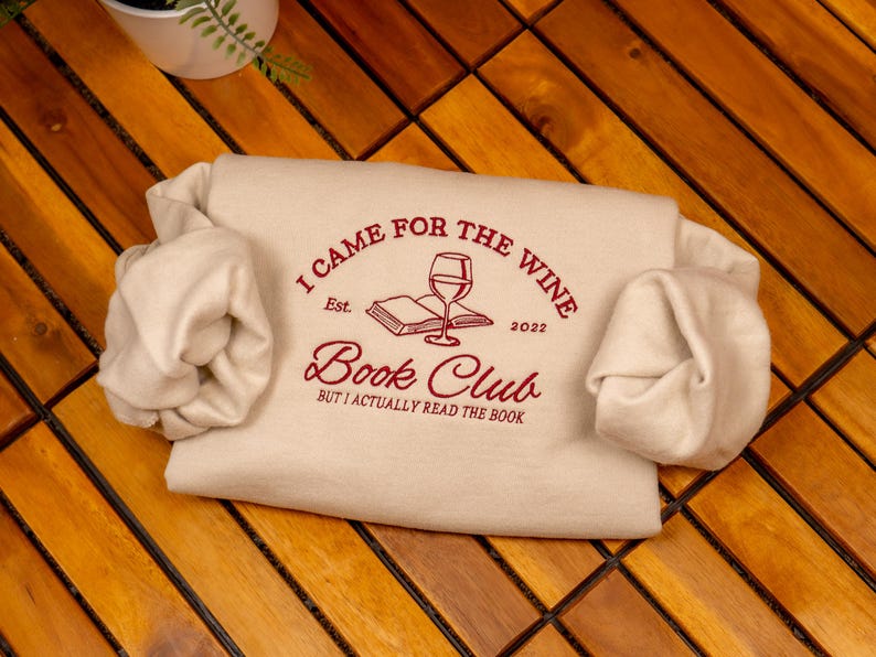 May include: A cream-colored sweatshirt featuring dark red embroidered text that reads I CAME FOR THE WINE, Est. 2022, Book Club, and BUT I ACTUALLY READ THE BOOK, along with a graphic illustration of an open book and a stemmed wine glass.