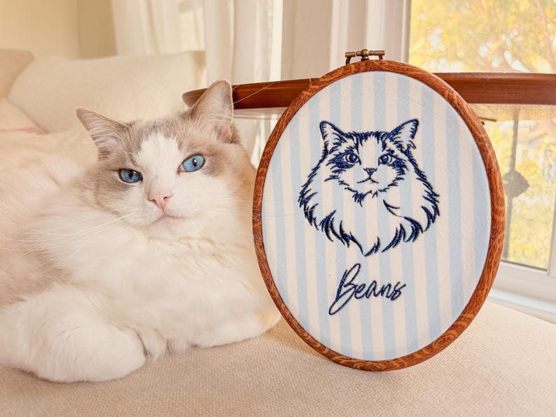 May include: An oval wooden embroidery hoop containing white fabric with light blue vertical stripes. The fabric features a dark blue embroidered portrait of a fluffy cat and the name Beans embroidered in a cursive script below the cat portrait.