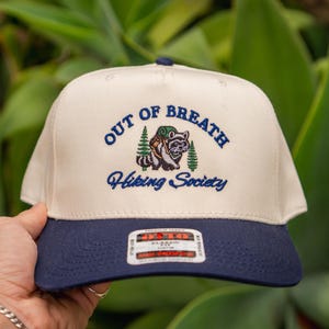 Op de afbeelding: Crème en marineblauwe baseballcap met geborduurde tekst en een wasbeer-afbeelding. De tekst luidt "OUT OF BREATH Hiking Society". De cap heeft een gebogen klep en een klein label.