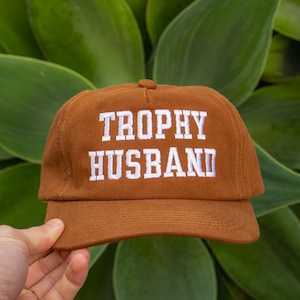 Puede incluir: Gorra de béisbol marrón con el texto bordado "TROPHY HUSBAND" en blanco. La gorra se sostiene frente a un fondo de hojas verdes.