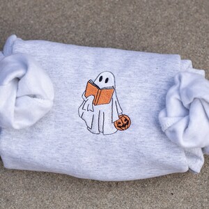 May include: A gray sweatshirt with a white ghost holding a book and a pumpkin. The ghost is embroidered on the sweatshirt.