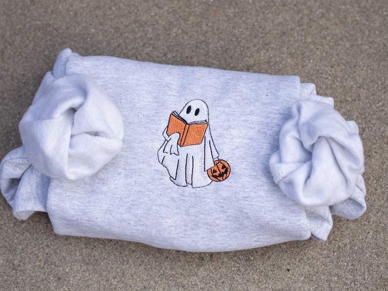 May include: A gray sweatshirt with a white ghost holding a book and a pumpkin. The ghost is embroidered on the sweatshirt.