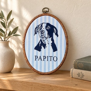 May include: An oval embroidery hoop frame containing fabric with light blue and white vertical stripes. The fabric features a dark blue embroidered portrait of a dog and the text PAPITO centered below the dog in a dark blue sans-serif font.