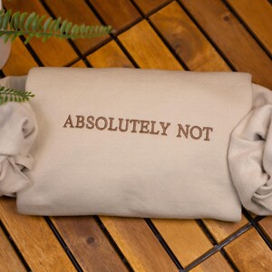 May include: A beige sweatshirt with the text "ABSOLUTELY NOT" embroidered in brown on the front. The sweatshirt is folded and lying on a wooden surface.