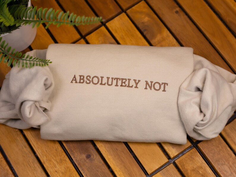 May include: A beige sweatshirt with the text "ABSOLUTELY NOT" embroidered in brown on the front. The sweatshirt is folded and lying on a wooden surface.