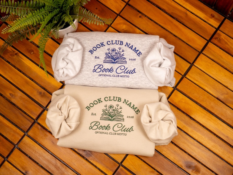 May include: Two folded sweatshirts, one light gray and one cream, each featuring a central graphic of an open book with flowers. The text reads "BOOK CLUB NAME", "Est. 2026", "Book Club", and "OPTIONAL CLUB MOTTO" in blue and dark green ink.
