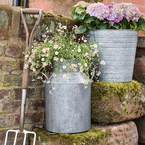 May include: A charming garden display with a metal milk churn and a galvanised bucket, both overflowing with flowers. A garden fork rests against a brick wall. The churn contains small white and pink flowers, the bucket, purple hydrangeas.