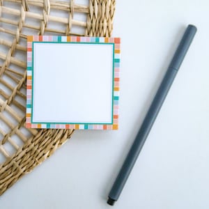 May include: A blank notepad with a colorful striped border sits on a woven wicker surface. A gray pen lies next to the notepad.