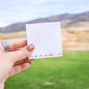 May include: A white notepad with a grid pattern and a rainbow border. The notepad is being held by a hand.