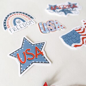 K&ouml;nnte beinhalten: Ein Set aus f&uuml;nf patriotischen Aufklebern in den Farben Rot, Wei&szlig; und Blau. Die Aufkleber beinhalten einen Regenbogen mit dem Wort "FREEDOM" und einem Herzen, einen Stern mit "USA" in roten Buchstaben, ein USA-Wortzeichen mit Sternen, ein USA-Wortzeichen mit Sternen und eine Karte der Vereinigten Staaten mit roten und wei&szlig;en Streifen.