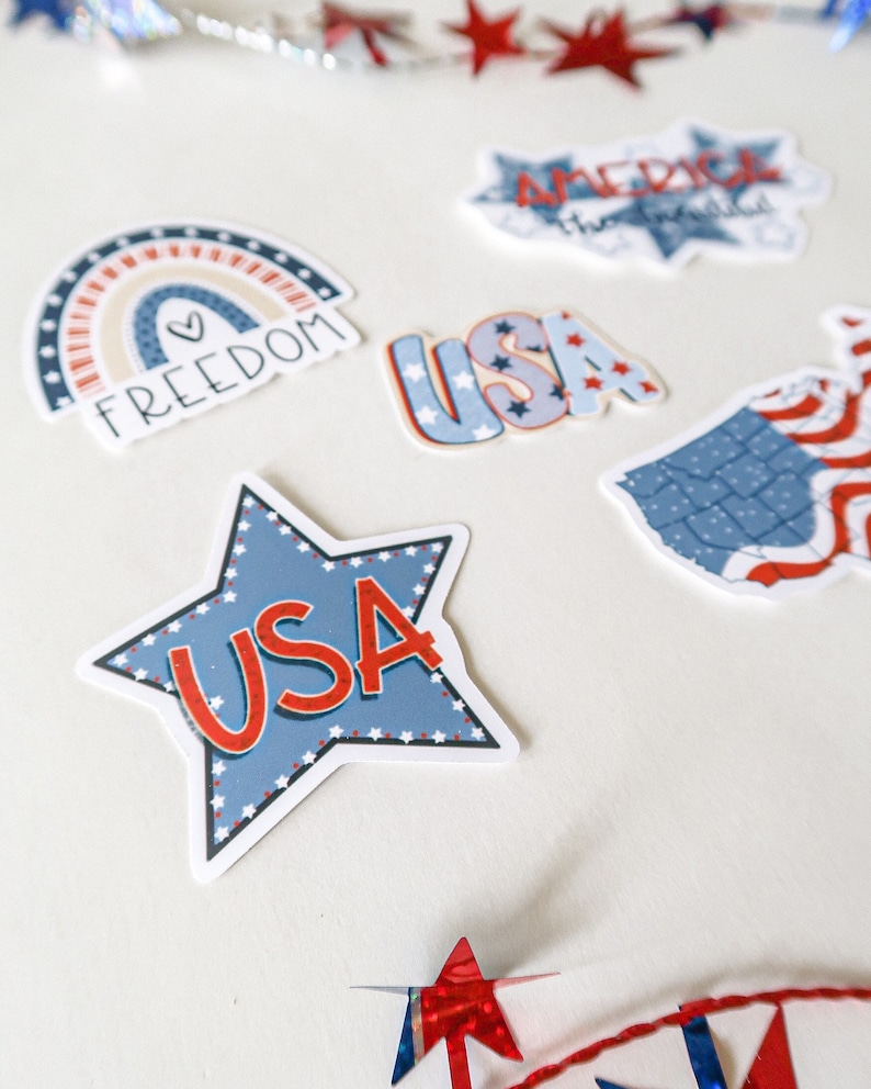 K&ouml;nnte beinhalten: Ein Set aus f&uuml;nf patriotischen Aufklebern in den Farben Rot, Wei&szlig; und Blau. Die Aufkleber beinhalten einen Regenbogen mit dem Wort "FREEDOM" und einem Herzen, einen Stern mit "USA" in roten Buchstaben, ein USA-Wortzeichen mit Sternen, ein USA-Wortzeichen mit Sternen und eine Karte der Vereinigten Staaten mit roten und wei&szlig;en Streifen.
