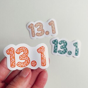 Puede incluir: Tres pegatinas con el texto "13.1" en un dise&ntilde;o floral. Las pegatinas son de color naranja, melocot&oacute;n y verde azulado. Las pegatinas est&aacute;n sobre un fondo blanco.