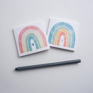 May include: Two rainbow-colored cards with a small heart in the center of each rainbow. The cards are on a white surface with a gray pen.