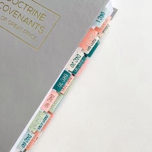 May include: A gray book with gold lettering and colorful page tabs. The tabs are labeled with dates like "DEC 10" and "DEC 20" in various colors. The book's title reads "THE DOCTRINE COVENANTS OF GREAT PRICE."