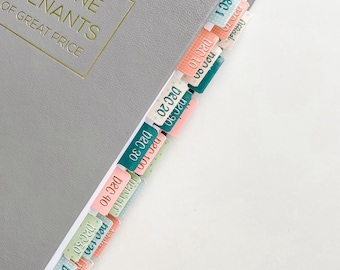 Puede incluir: Un libro gris con letras doradas y pestañas de página coloridas. Las pestañas están etiquetadas con fechas como "DEC 10" y "DEC 20" en varios colores. El título del libro dice "THE DOCTRINE COVENANTS OF GREAT PRICE."