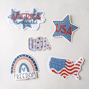 K&ouml;nnte beinhalten: F&uuml;nf patriotische Aufkleber in den Farben Rot, Wei&szlig; und Blau. Die Aufkleber zeigen einen Regenbogen mit dem Wort "FREEDOM", einen Stern mit "USA" darin, eine Karte der Vereinigten Staaten mit einem amerikanischen Flaggenmuster und zwei verschiedene Designs, die "USA" und "AMERICA the beautiful" sagen.