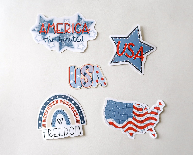 K&ouml;nnte beinhalten: F&uuml;nf patriotische Aufkleber in den Farben Rot, Wei&szlig; und Blau. Die Aufkleber zeigen einen Regenbogen mit dem Wort "FREEDOM", einen Stern mit "USA" darin, eine Karte der Vereinigten Staaten mit einem amerikanischen Flaggenmuster und zwei verschiedene Designs, die "USA" und "AMERICA the beautiful" sagen.