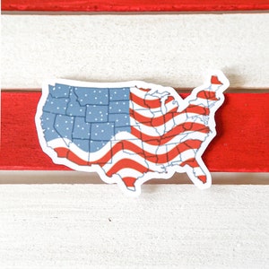 K&ouml;nnte beinhalten: Ein Aufkleber der Vereinigten Staaten von Amerika in Form des Landes mit dem Design der amerikanischen Flagge. Der Aufkleber befindet sich auf einem rot-wei&szlig; gestreiften Hintergrund.