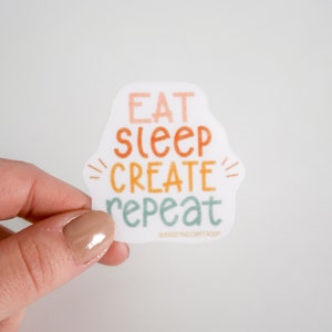 May include: A white sticker with the text "Eat Sleep Create Repeat" in a colorful font. The text is arranged in a vertical stack with the words "Create" in a larger font size.