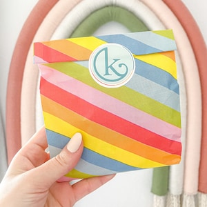 May include: A colorful package with diagonal rainbow stripes is held in a hand. The package has a circular sticker with a teal "K" logo. A rainbow wall hanging is in the background.