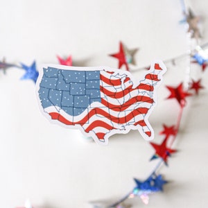K&ouml;nnte beinhalten: Ein wei&szlig;er Aufkleber der Vereinigten Staaten von Amerika mit einem rot-wei&szlig;-blauen amerikanischen Flaggenmuster. Der Aufkleber befindet sich vor einem wei&szlig;en Hintergrund mit einer verschwommenen Girlande aus rot-wei&szlig;-blauen Sternen.