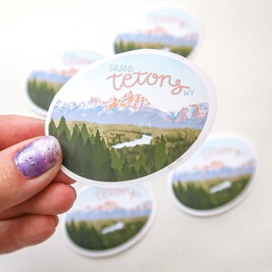 Pode incluir: Um adesivo redondo com uma imagem c&ecirc;nica das montanhas Grand Teton em Wyoming. O adesivo apresenta um c&eacute;u azul, &aacute;rvores verdes e um lago em primeiro plano. O texto "GRAND TETONS WY" est&aacute; impresso em rosa no adesivo.
