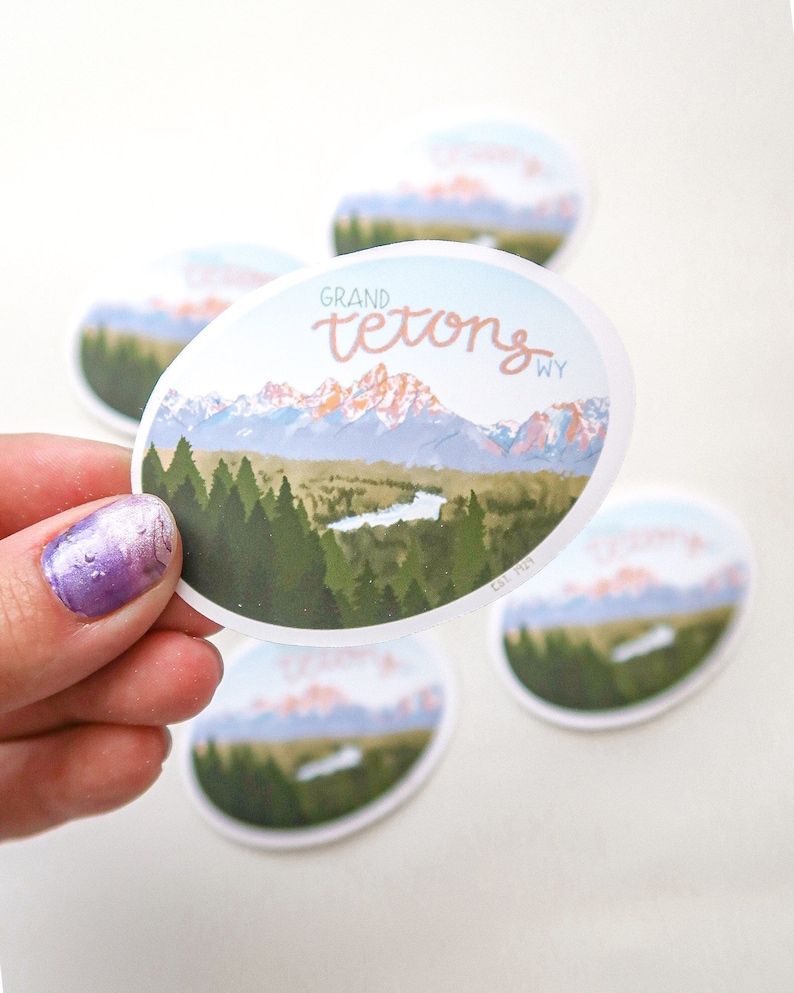 Pode incluir: Um adesivo redondo com uma imagem c&ecirc;nica das montanhas Grand Teton em Wyoming. O adesivo apresenta um c&eacute;u azul, &aacute;rvores verdes e um lago em primeiro plano. O texto "GRAND TETONS WY" est&aacute; impresso em rosa no adesivo.