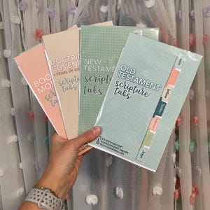 May include: A hand holds a stack of scripture tabs in various pastel colors. The tabs are labeled "Book of Mormon," "Doctrine & Covenants," "New Testament," and "Old Testament." Each tab set has a different background color and the text is in a handwritten style font.