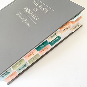May include: A gray "The Book of Mormon Journal Edition" book with colorful tabs. The tabs are labeled with names like "Alma", "Jacob", and "Moroni". The book is open, revealing the tabs.