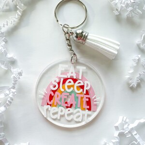 May include: A white acrylic keychain with a rainbow design and the text "Eat Sleep Create Repeat". The keychain has a silver key ring and a white tassel.