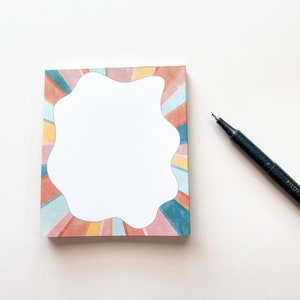 May include: A notepad with a colorful abstract design featuring a white, irregular-shaped space in the center. The notepad is on a white surface with a black pen.
