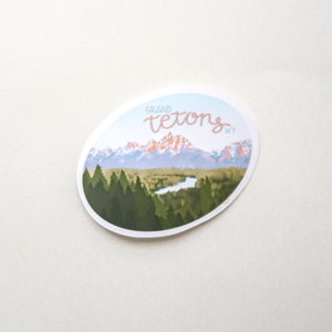 Pode incluir: Um adesivo com uma vista panor&acirc;mica das montanhas Grand Teton em Wyoming. O adesivo apresenta um c&eacute;u azul, montanhas cobertas de neve, um rio e uma floresta de &aacute;rvores verdes. O texto "GRAND TETONS WY" est&aacute; escrito em it&aacute;lico rosa.