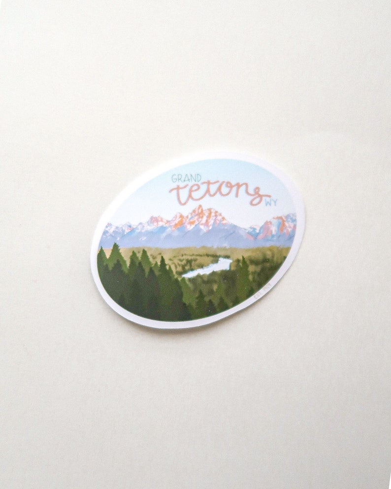 Pode incluir: Um adesivo com uma vista panor&acirc;mica das montanhas Grand Teton em Wyoming. O adesivo apresenta um c&eacute;u azul, montanhas cobertas de neve, um rio e uma floresta de &aacute;rvores verdes. O texto "GRAND TETONS WY" est&aacute; escrito em it&aacute;lico rosa.