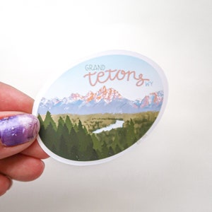 Pode incluir: Um adesivo redondo com uma vista panor&acirc;mica da cordilheira das montanhas Grand Teton em Wyoming. O adesivo apresenta um c&eacute;u azul, montanhas cobertas de neve, um rio e &aacute;rvores verdes. O texto "GRAND TETONS WY" est&aacute; escrito em uma fonte cursiva.