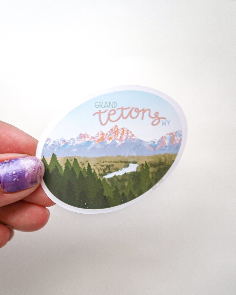 Pode incluir: Um adesivo redondo com uma vista panor&acirc;mica da cordilheira das montanhas Grand Teton em Wyoming. O adesivo apresenta um c&eacute;u azul, montanhas cobertas de neve, um rio e &aacute;rvores verdes. O texto "GRAND TETONS WY" est&aacute; escrito em uma fonte cursiva.
