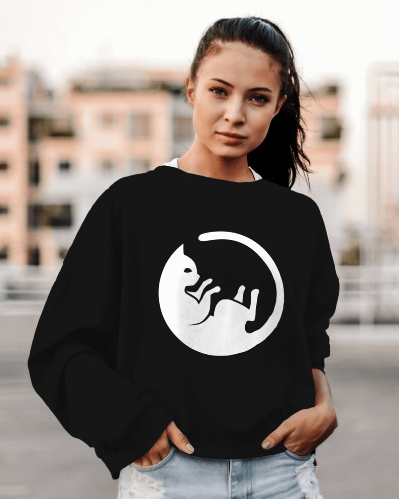 May include: A black sweatshirt featuring a white graphic of a cat curled up in a circle. The cat is facing to the left and its tail is curled around its body.