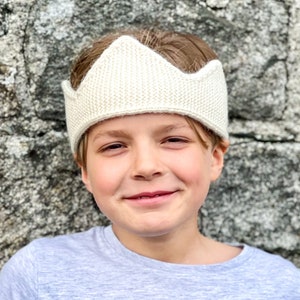 Könnte beinhalten: Ein junges Kind trägt ein weißes, gestricktes Stirnband in Form einer Krone. Das Stirnband hat eine spitze Kronenform und ist aus weichem, strukturiertem Garn gefertigt.