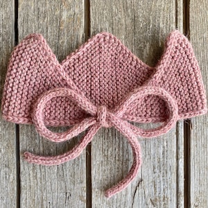 May include: A pink knitted crown headband with a bow tied in the front.