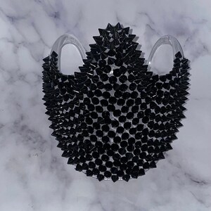 May include: A black face mask with a unique design featuring hundreds of small black spikes. The mask is made of a clear material and has two loops for the ears.