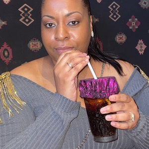 Op de afbeelding: Een vrouw draagt een grijze trui met gouden franjes op één schouder en houdt een roze en zwarte herbruikbare beker vast met een rietje. Ze zit voor een gepatroneerde achtergrond.