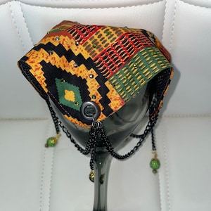 Puede incluir: Un tocado decorativo con un estampado africano vibrante en amarillo, negro, rojo y verde, adornado con pedrería. Cadenas negras y detalles de cuentas verdes cuelgan de la parte delantera. El tocado se muestra en una copa de vino gris oscuro.